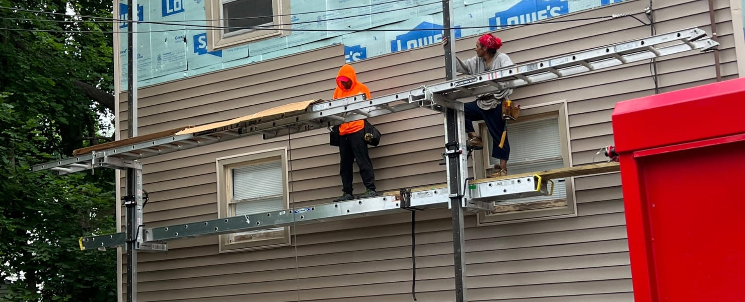 Active residential siding replacement in Albany, NY, featuring contractors on pump jack scaffolding installing tan vinyl panels over Lowe's GreenGuard moisture barrier by MG Contractor NY