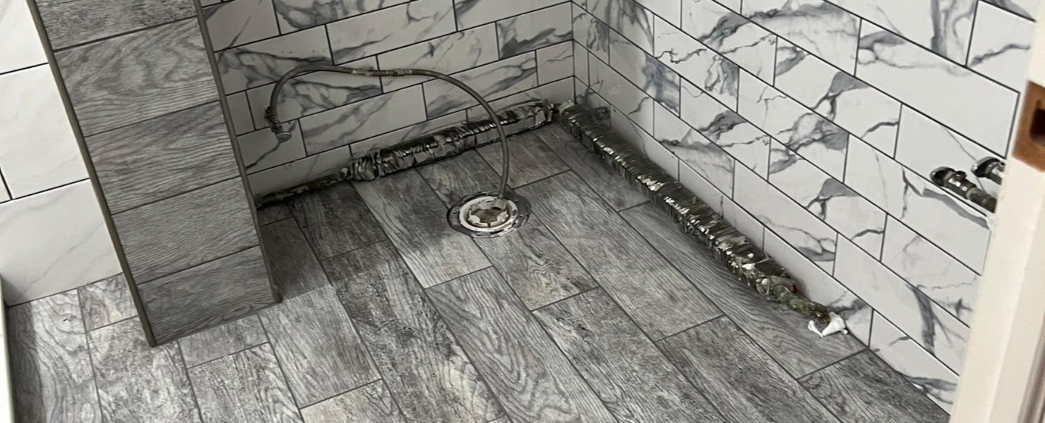 n-progress bathroom renovation showing installation of white marble subway tiles and gray wood-grain floor tiles with exposed plumbing for a modern custom shower